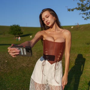 A woman in a strapless brown corset and a white lace skirt is taking a selfie with a smartphone in a grassy field.