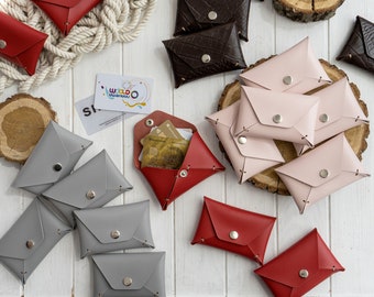 May include: Assortment of small leather wallets in various colors, including gray, pink, red, and brown. The wallets are all closed and have a single button closure. Some of the wallets have a textured surface. The wallets are arranged on a white wooden surface.