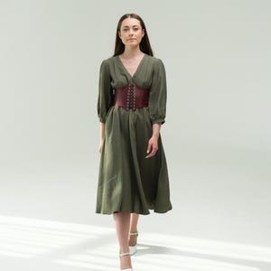 A woman wearing a green dress with a red belt walks confidently in front of a plain white background.