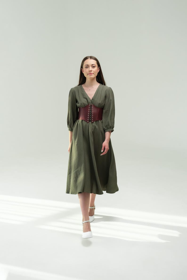 A woman wearing a green dress with a red belt walks confidently in front of a plain white background.