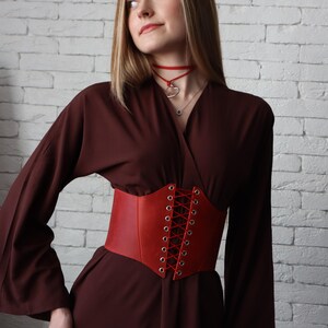 A woman in a brown dress with a red corset stands against a white brick wall, her gaze directed upwards.