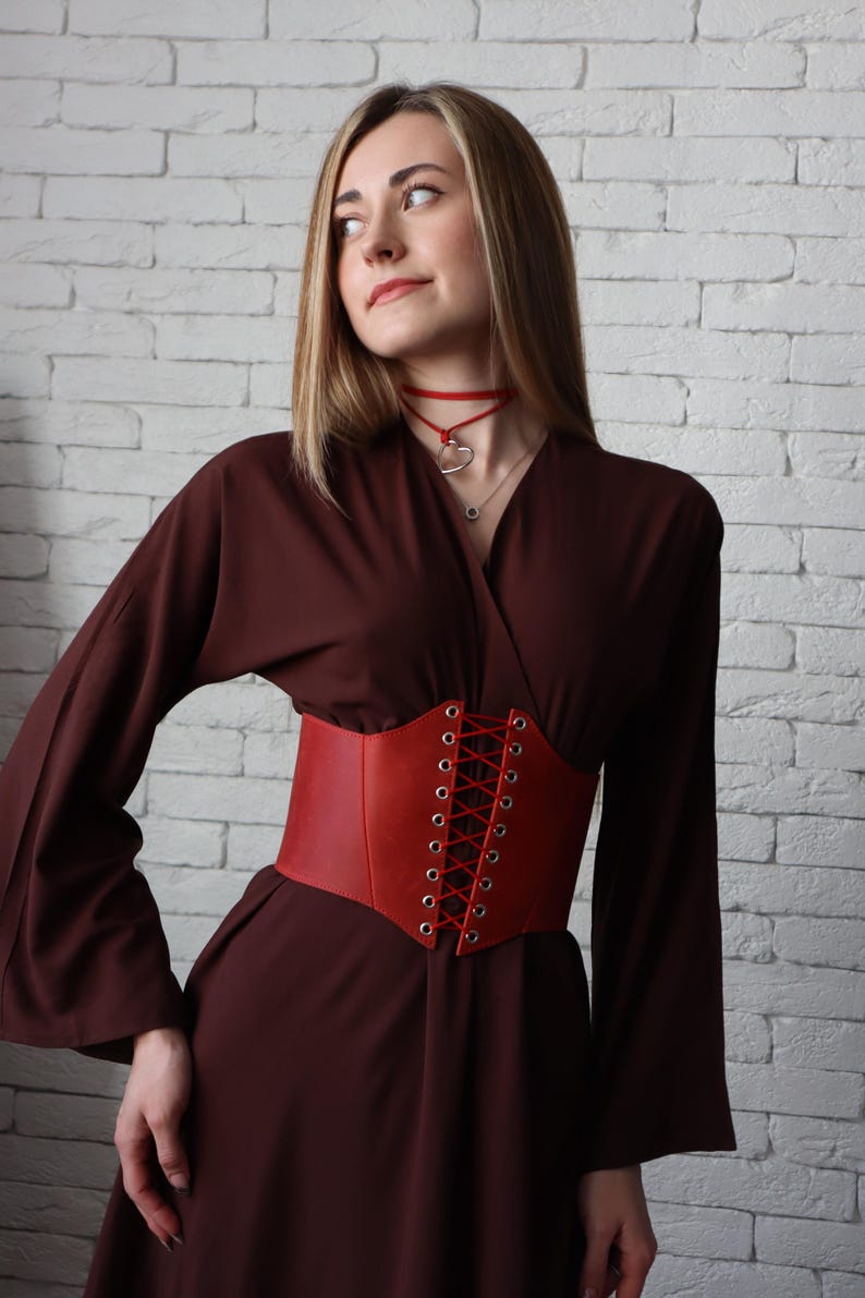 A woman in a brown dress with a red corset stands against a white brick wall, her gaze directed upwards.