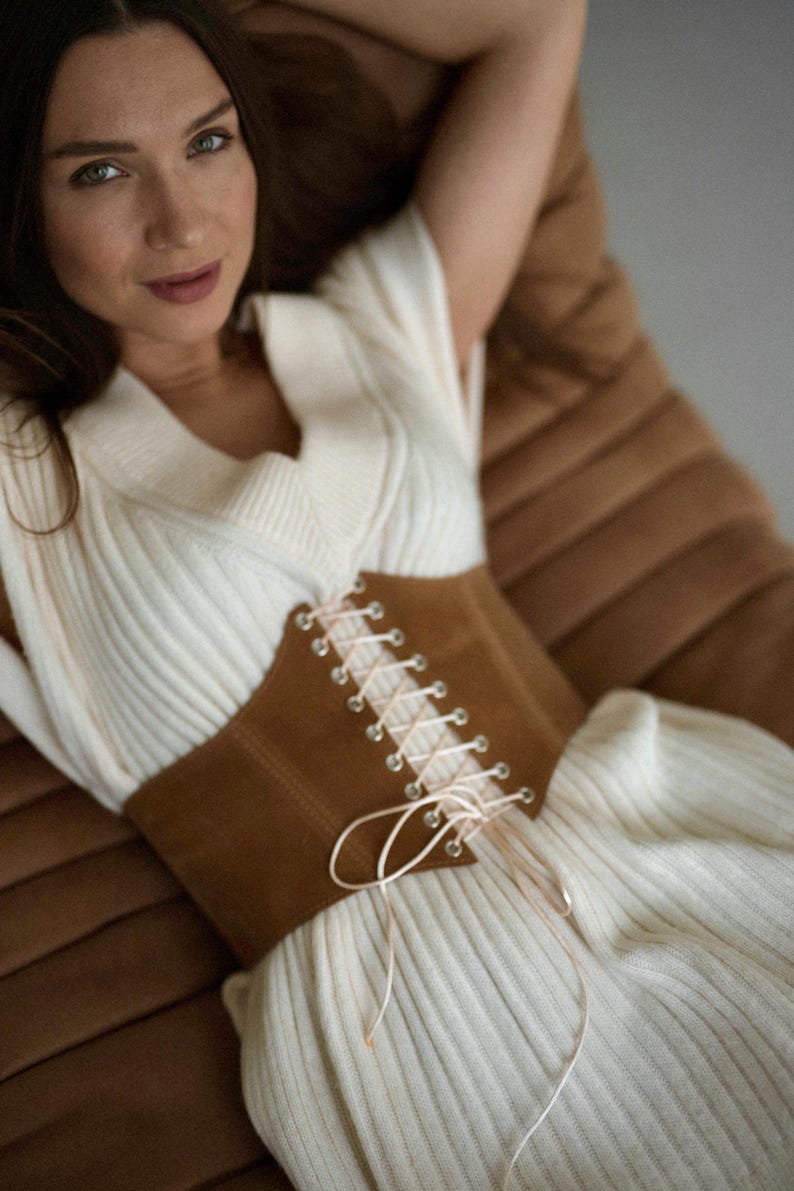 A woman wearing a white sweater with a brown corset sits on a brown leather couch, looking directly at the camera with a relaxed and confident expression.