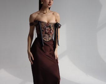 May include: A woman's outfit comprising a floral corset with black ribbon detailing and a long, dark brown satin skirt. The ensemble is complemented by black boots. Gold jewellery adds a touch of elegance, creating a sophisticated look.