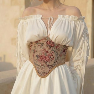 May include: A white off-the-shoulder dress with a floral tapestry corset. The corset features a beige background with red and pink floral patterns. The dress has long sleeves with drawstring details and a V-neckline.
