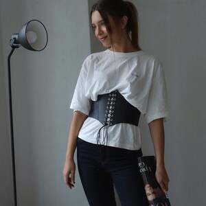 A woman wearing a white shirt and black pants stands in a room, holding a magazine and looking to her left.