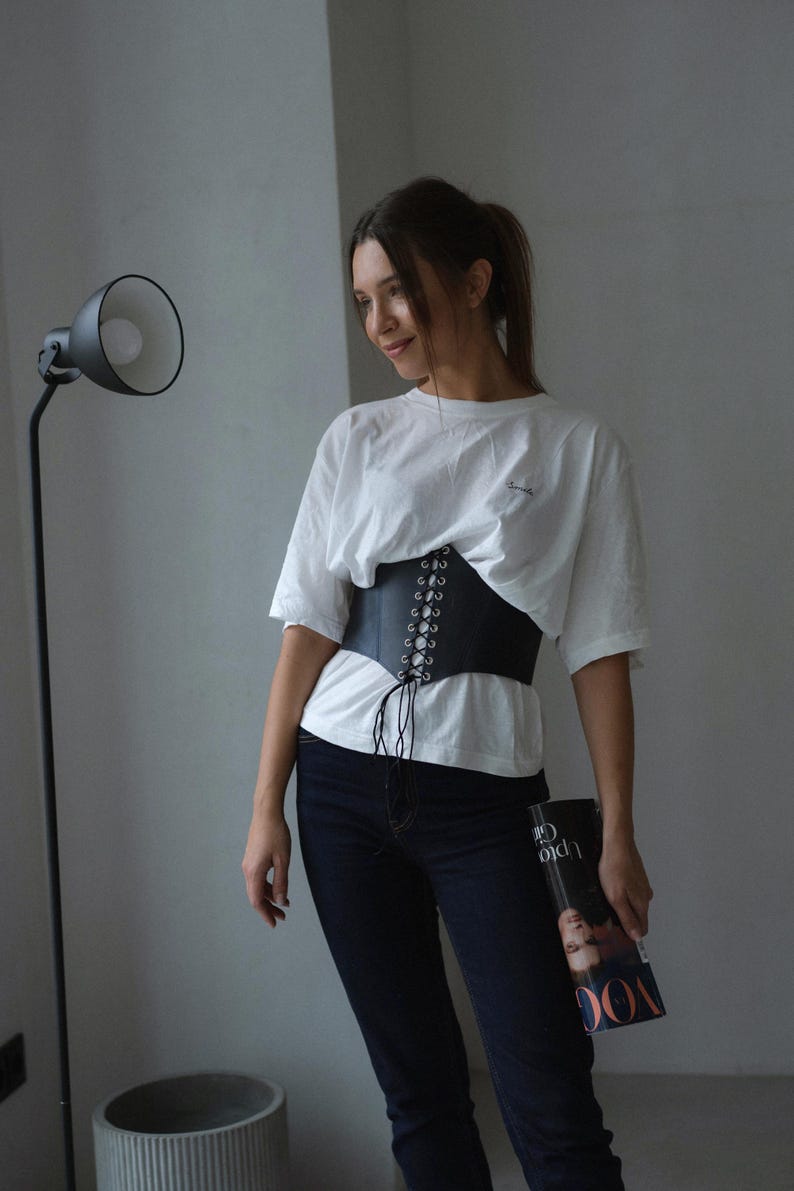 A woman wearing a white shirt and black pants stands in a room, holding a magazine and looking to her left.