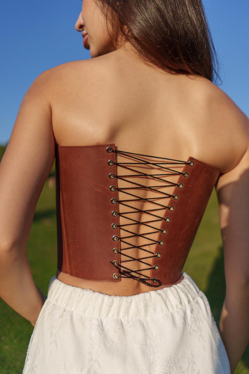 A woman wearing a brown leather corset with a lace-up back, standing in a grassy field.