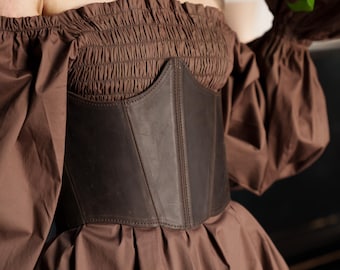 May include: A brown off-the-shoulder dress with a dark brown leather corset. The dress features a gathered bodice and long, puffed sleeves. The corset is stitched and fitted, adding a structured element to the garment.