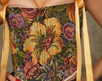 May include: A floral tapestry corset top with yellow ribbon straps. The corset features a vibrant pattern of large yellow and orange flowers, with accents of purple, red, and green. The straps are tied with long, flowing yellow ribbons.