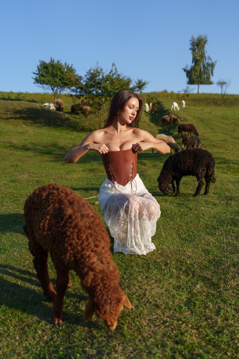 A woman in a brown dress stands in a grassy field, surrounded by a group of sheep grazing in the background.