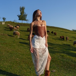 May include: A brown leather corset with lace-up detailing is paired with a white lace skirt and brown boots. The model is standing on a grassy hill with sheep grazing in the background under a clear blue sky.