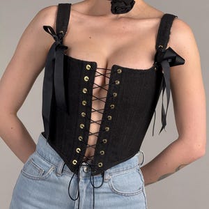May include: Black denim corset with gold-tone eyelets and black ribbon lacing. The corset features shoulder straps with black bows. A black choker with a rose detail is also visible. The corset is paired with light blue denim jeans.
