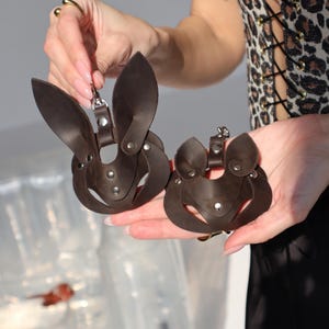 May include: Two brown leather bunny masks with metal accents. The masks are held in the hands of a person wearing a leopard print corset.