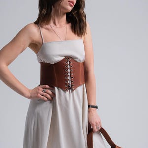 A woman in a white dress with a brown belt stands against a plain background, holding a brown purse.