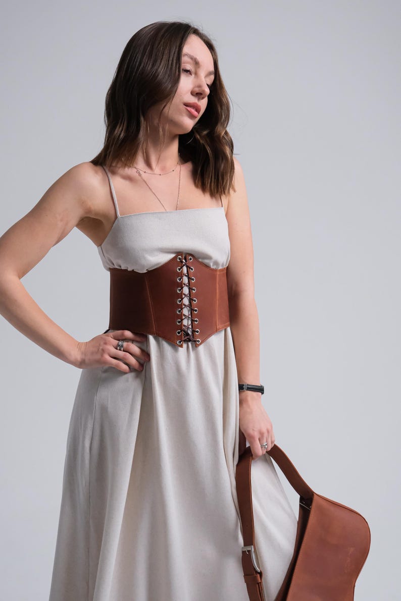 A woman in a white dress with a brown belt stands against a plain background, holding a brown purse.