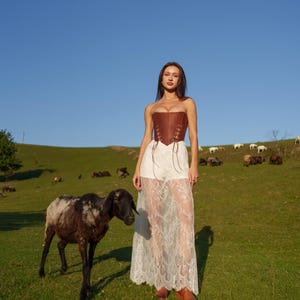 A woman in a white lace dress stands in a grassy field, accompanied by a black and white sheep.