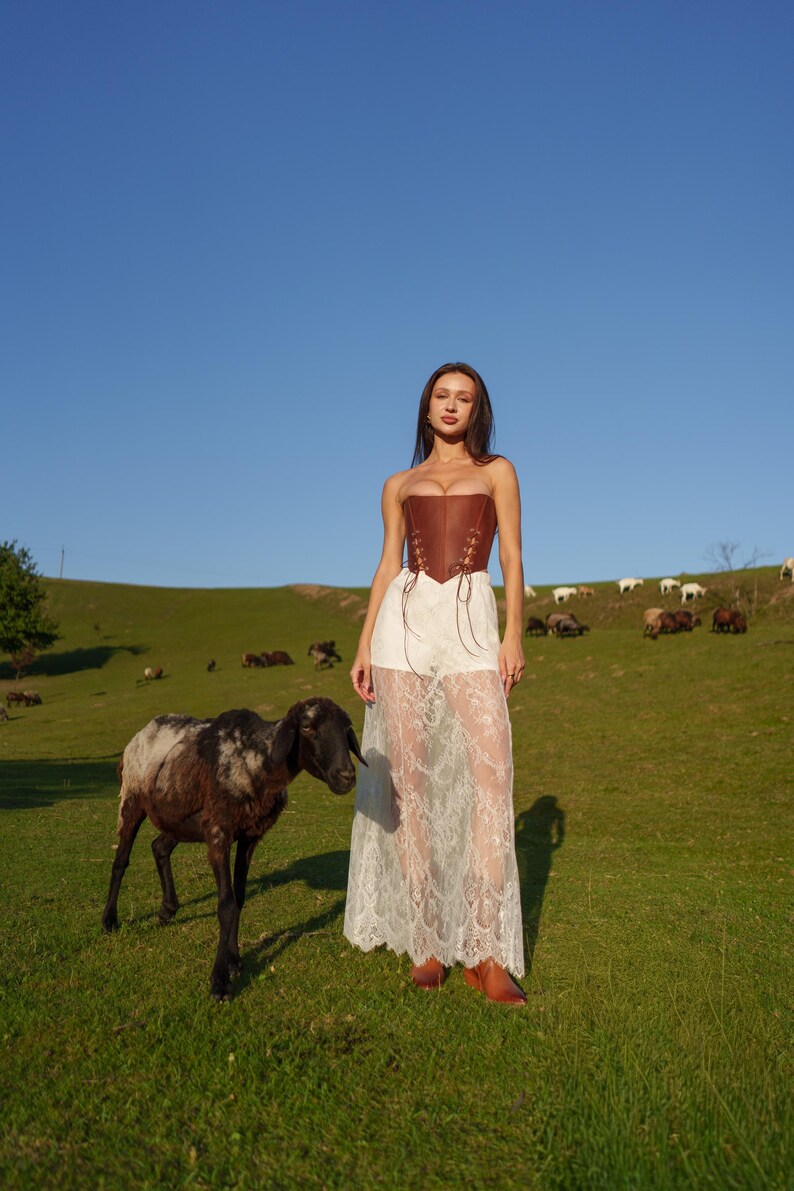 A woman in a white lace dress stands in a grassy field, accompanied by a black and white sheep.