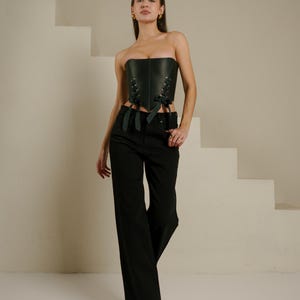 A woman wearing a black strapless corset and wide-leg pants stands in front of a white staircase, posing confidently.
