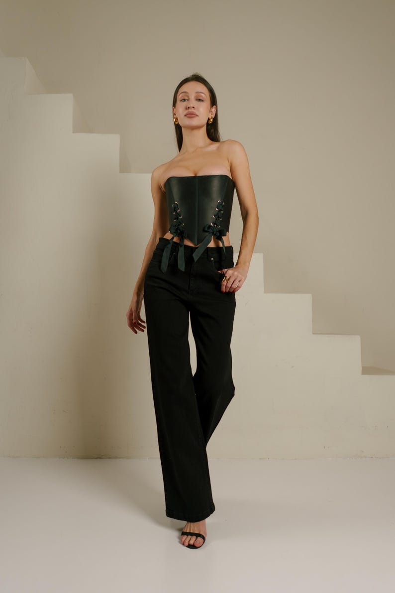A woman wearing a black strapless corset and wide-leg pants stands in front of a white staircase, posing confidently.