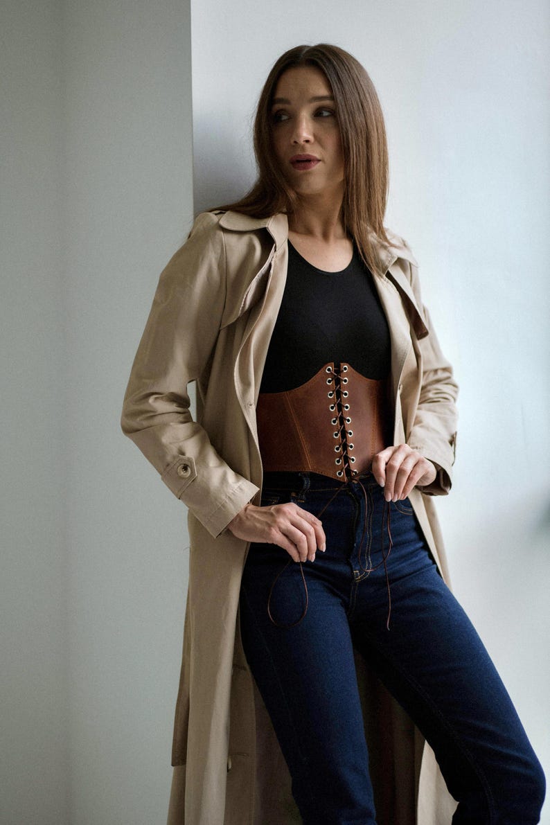 A woman wearing a beige trench coat, black top, and blue jeans stands against a white wall, looking to her left with a thoughtful expression.