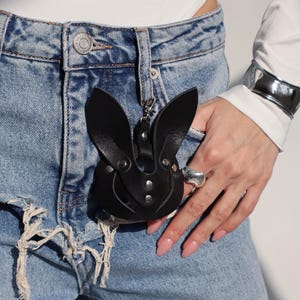 May include: A black leather bunny-shaped keychain with a silver ring attached. The keychain is hanging from the front pocket of a pair of blue denim jeans.