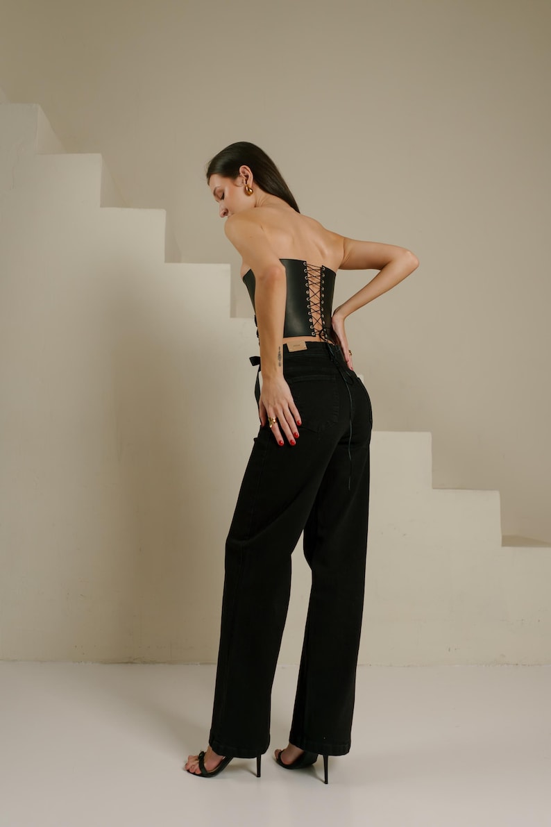 A woman wearing a strapless black corset and wide-leg pants stands in front of a white staircase, with her back turned to the camera.