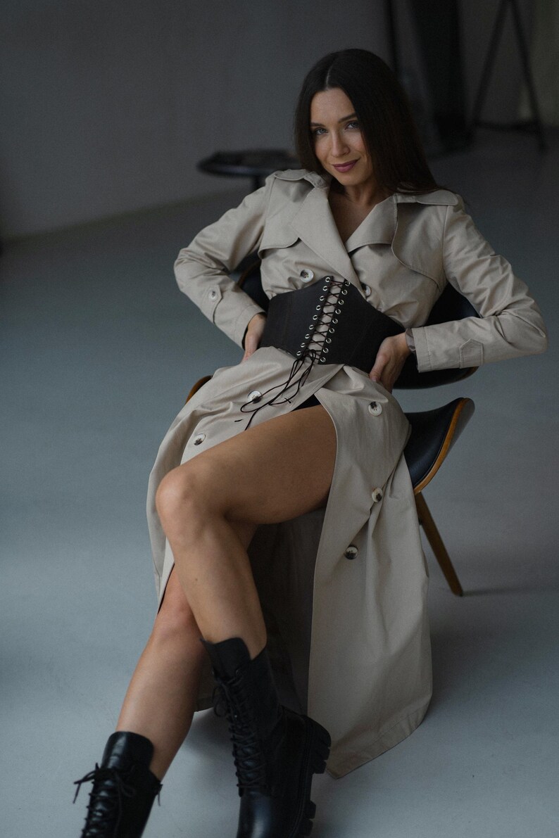 A woman in a beige trench coat and black boots sits on a chair, smiling at the camera.
