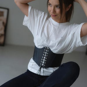 A woman in a white shirt and black corset sits on a stool, her hands on her head, with a thoughtful expression on her face.