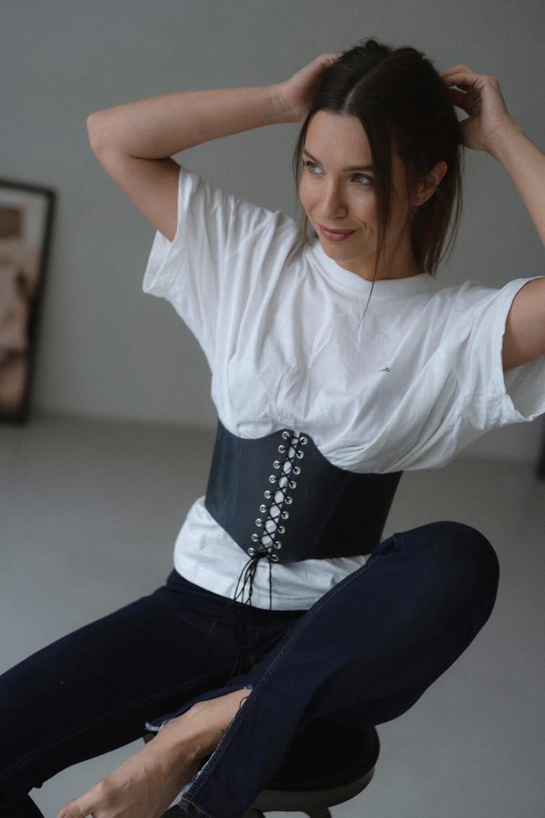 A woman in a white shirt and black corset sits on a stool, her hands on her head, with a thoughtful expression on her face.