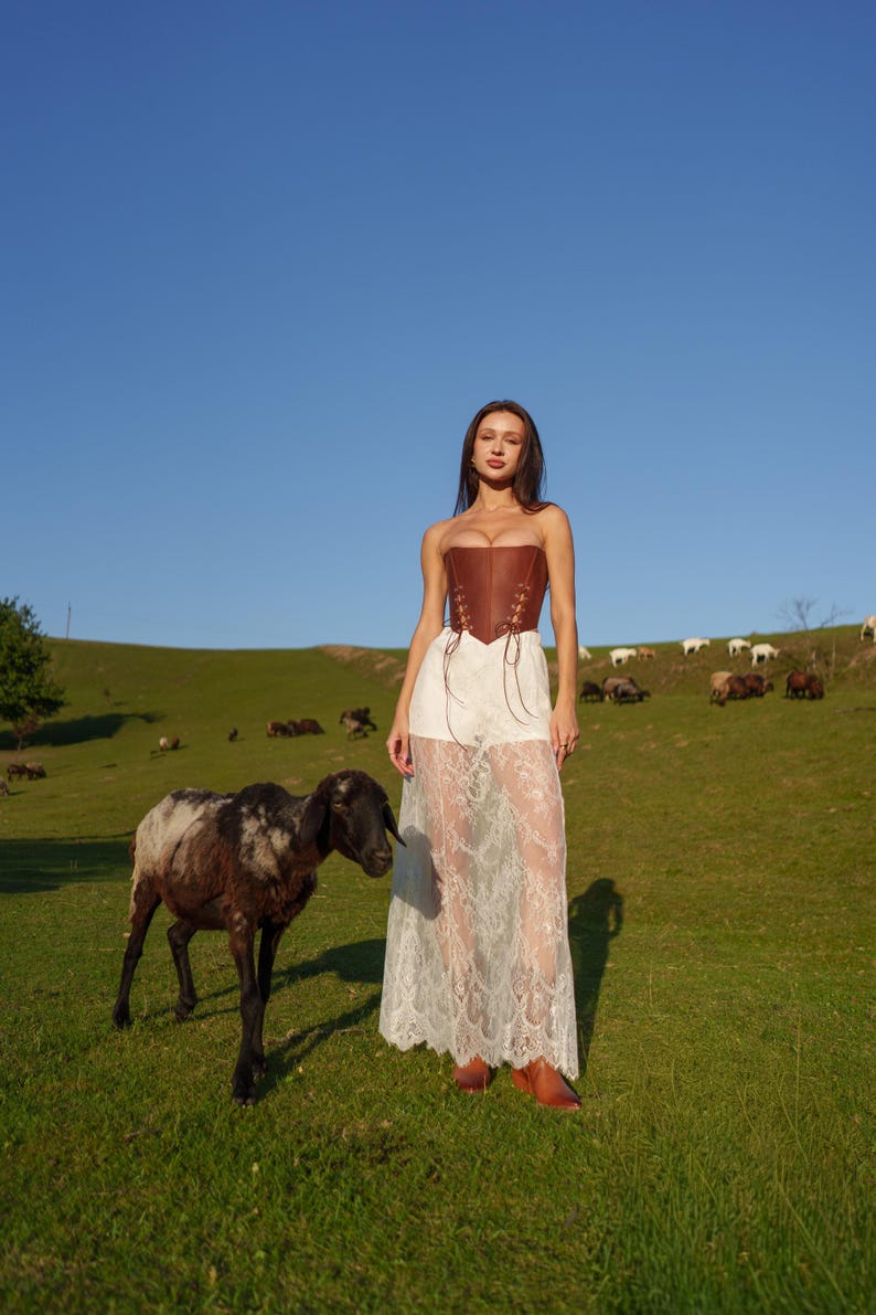 A woman in a white lace dress stands in a grassy field, accompanied by a black and white sheep.