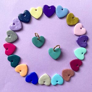 Pode incluir: Uma coleção de contas coloridas em forma de coração e dois brincos de coração turquesa com argolas douradas, dispostos sobre uma superfície roxa. As contas são de várias cores, incluindo rosa, azul, amarelo e roxo. Os brincos estão no centro.