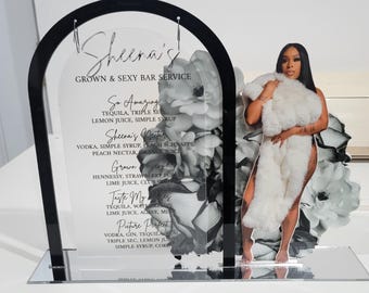 May include: A black acrylic arch with a clear panel featuring a cocktail menu with the title "Sheena's Grown & Sexy Bar Service". The menu lists cocktails with names like "So Amazing", "Sheena's Nectar", "Grown Gay", "Taste My", and "Picture Perfect". The arch is decorated with a black and white floral design and a cutout of a woman in a white dress.