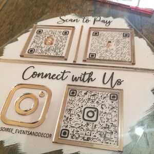 Pode incluir: Uma placa branca com bordas douradas que apresenta quatro c&oacute;digos QR. Os dois c&oacute;digos QR superiores s&atilde;o etiquetados como "Scan to Pay". Os dois c&oacute;digos QR inferiores s&atilde;o etiquetados como "Connect with Us". O c&oacute;digo QR inferior esquerdo est&aacute; ao lado de um logotipo do Instagram. A placa tem o texto "soiree_eventsanddecor" na parte inferior.