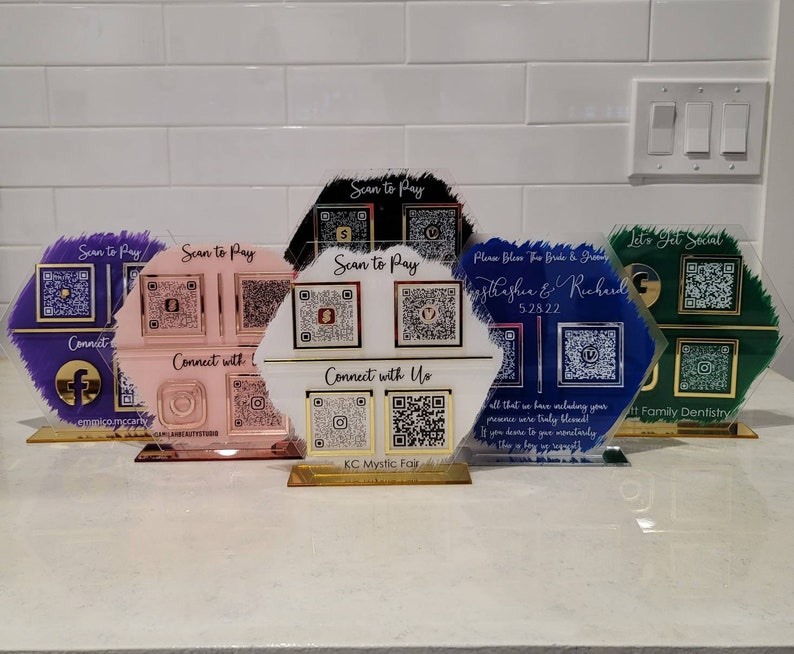 Pode incluir: Cinco placas hexagonais de acr&iacute;lico com bordas douradas e uma base dourada escovada. Cada placa tem um fundo de cor diferente e apresenta um c&oacute;digo QR e texto. O texto nas placas inclui "Scan to Pay", "Connect with Us", "Let's Get Social", "Please Bless The Bride & Groom", e "KC Mystic Fair".