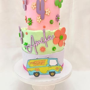 May include: A three-tiered birthday cake with pink, green, and lavender frosting. The top tier has a purple number 4 and flower decorations. The middle tier has the name "Amelia" and orange flowers. The bottom tier features a cartoon image of "The Mystery Machine" van.