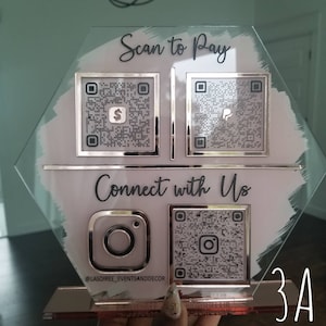 Pode incluir: Um sinal de vidro transparente com fundo branco e texto preto. O sinal tem quatro c&oacute;digos QR, um para cada um dos seguintes: Scan to Pay, Connect with Us, Instagram e um c&oacute;digo QR gen&eacute;rico. O texto "@lasoiree_eventsanddecor" &eacute; impresso abaixo do c&oacute;digo QR do Instagram. O sinal tamb&eacute;m tem o texto "Scan to Pay" e "Connect with Us" impresso nele. O sinal est&aacute; sobre uma superf&iacute;cie branca com um fundo rosa e branco.