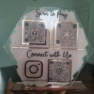 Pode incluir: Uma placa de vidro com fundo branco e texto preto que diz "Scan to Pay" e "Connect with Us". Existem quatro c&oacute;digos QR na placa, dois para pagamento e dois para m&iacute;dia social. A placa tamb&eacute;m inclui o logotipo do Instagram e o texto "@EVENTIENDDETS&T3DECOR".
