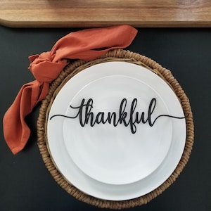 May include: A white plate with a black wooden 'thankful' sign on top of it. The plate is on a woven placemat with a burnt orange napkin.