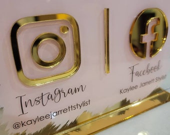 May include: A pink and gold sign with the text "Connect with Me" and social media icons for Instagram and Facebook. The Instagram icon is a gold outline of the Instagram logo with a pink background. The Facebook icon is a gold outline of the Facebook logo with a pink background. The text "Instagram" is below the Instagram icon and the text "@kayleejarrettstylist" is below that. The text "Facebook" is below the Facebook icon and the text "Kaylee Jarrett Stylist" is below that.