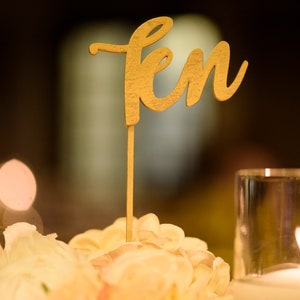 May include: A gold wooden table number with the word "ten" written in cursive script. The number is placed on a table setting with white flowers and a candle.