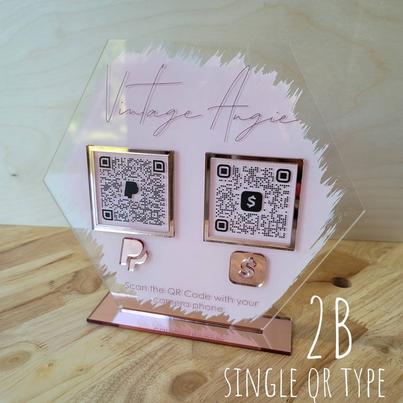 Pode incluir: Um sinal hexagonal de acr&iacute;lico transparente com fundo rosa e detalhes em ouro rosa. O sinal apresenta dois c&oacute;digos QR, um para PayPal e outro para Square, com os logotipos correspondentes abaixo. O texto "Vintage Angie" est&aacute; escrito em letra de ouro rosa na parte superior do sinal. O texto "Digitalize o c&oacute;digo QR com seu telefone" est&aacute; escrito abaixo dos c&oacute;digos QR. O sinal est&aacute; em um suporte de ouro rosa com o texto "2B SINGLE QR TYPE" escrito abaixo do suporte.
