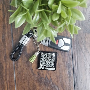 May include: Black and white QR code keychain with a green tassel and a black leather keychain with the Audi logo. The keychain is on a wooden surface.