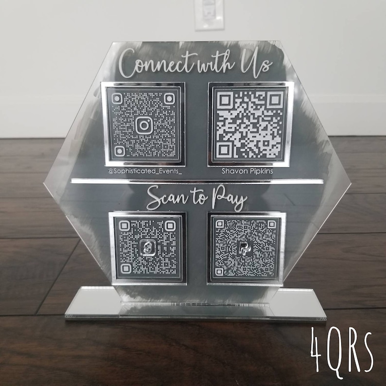 Pode incluir: Um sinal hexagonal de acr&iacute;lico com fundo cinza e letras prateadas. O sinal tem quatro c&oacute;digos QR, dois etiquetados como "@Sophisticated_Events_" e "Shavon Pipkins" e dois etiquetados como "Scan to Pay". O sinal est&aacute; em um suporte espelhado com o texto "4QRS" impresso na base.