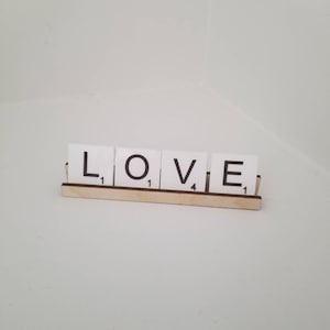 May include: White Scrabble tiles spelling out the word "LOVE" on a wooden rack. The tiles have the letter values printed on them.