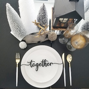 May include: A white plate with a black 'together' sign on a black table setting with gold flatware, a gold deer figurine, and white pine trees.