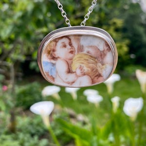 Puede incluir: Collar con colgante plateado con un camafeo ovalado de porcelana con una escena de querubines pintados. El colgante cuelga de una delicada cadena. El fondo es una vista borrosa de vegetación y flores blancas.