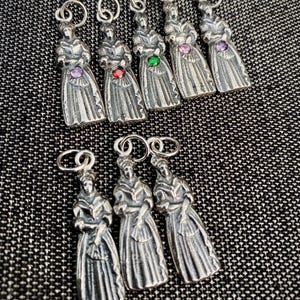 Puede incluir: Un conjunto de ocho dijes de metal en tono plateado, cada uno con forma de figura con vestido largo. Cinco de los dijes presentan una pequeña piedra preciosa redonda y facetada en colores morado, rojo, verde y rosa, mientras tres son lisos.