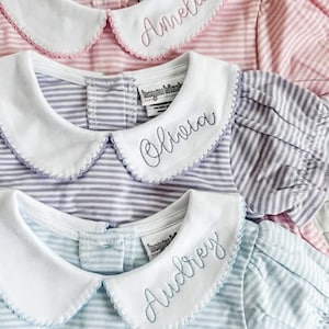 Puede incluir: Tres vestidos de bebé a rayas en rosa, lavanda y azul. Cada vestido tiene un cuello Peter Pan blanco con un borde festoneado y un nombre bordado en cursiva: Amelia, Olivia y Audrey. Los vestidos tienen mangas cortas abullonadas.