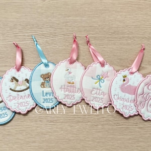 May include: Embroidered baby gift tags with various designs, including a rocking horse, teddy bear, and swan. Each tag has a name and the year 2025, attached with a pink or blue ribbon. The tags have a scalloped edge.